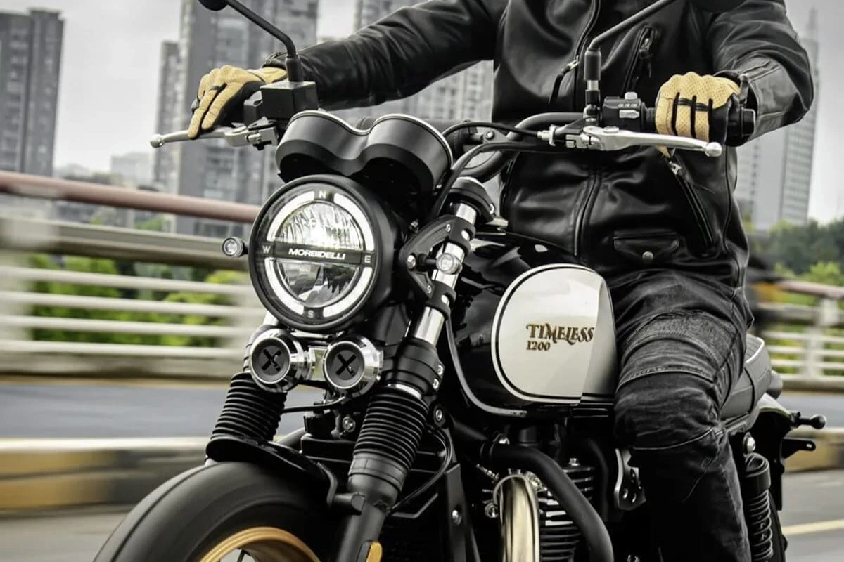 Le phare, le réservoir et la partie avant d'une moto Morbidelli 1200 Timeless, des gants de moto beiges et un blouson moto noir.