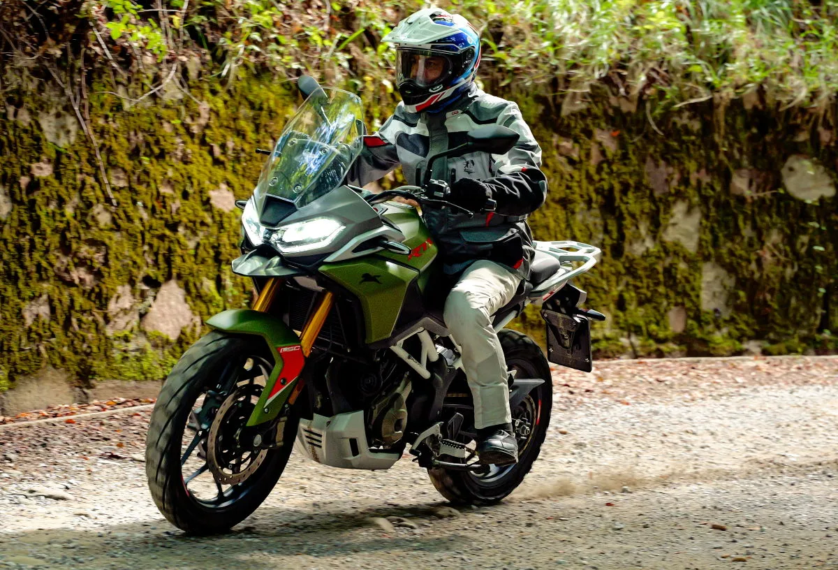 Une moto TVS RTX 300 sur un chemin, un motard en tenue trail grise et un casque bleu blanc rouge Shoei Hornet.