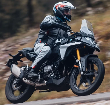 Una moto BMW F 450 GS negra y gris, un paisaje rocoso difuminado, un motorista vestido con un conjunto de chaqueta y cazadora de tela estilo trail-aventura y un casco crossover Shoei azul, blanco y rojo