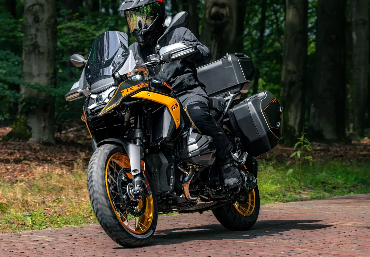 Une moto BMW R 1300 GS Aurum Edition jaune avec des valises et un top-case roulant sur des pavés et une forêt.