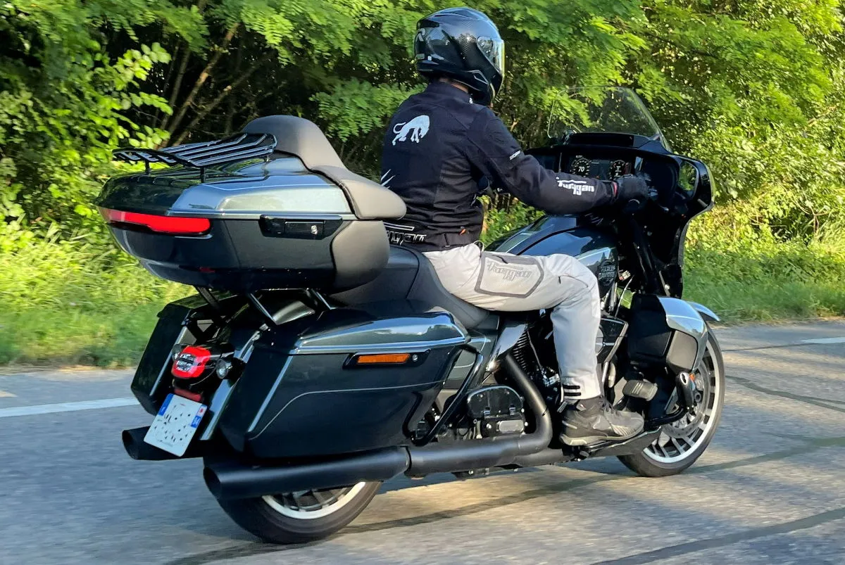 Une moto Harley-Davidson Street Glide Limited sur la route, un motard en pantalon blanc et blouson noir, des arbres.