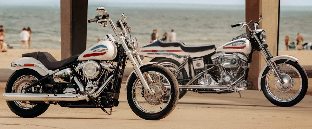 Une moto Harley-Davidson 1920 Super Glide FXD blanche, une Superglide de 1971, une plage, l'océan et deux piliers.