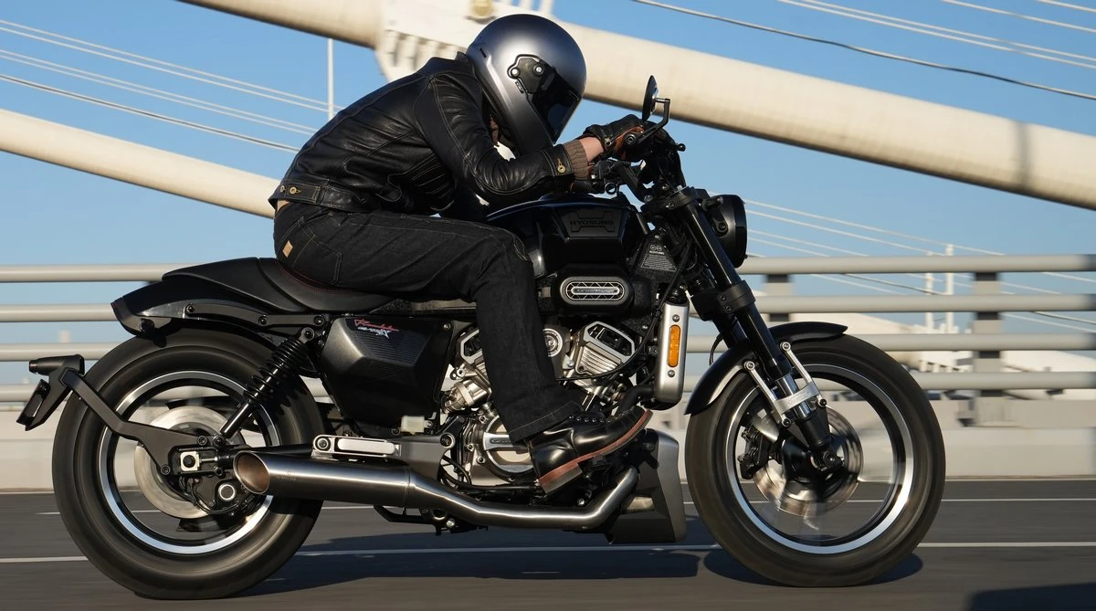 Une moto Hyosung GV125X noire, un motard habillé en noir, un ciel bleu et la structure d'un pont