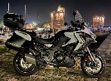 Une moto Kawasaki Versys 1100 SE blanche avec des valises et un top-case garée devant le port de La Rochelle, avec deux vieilles tours en pierre illuminées en pleine nuit.