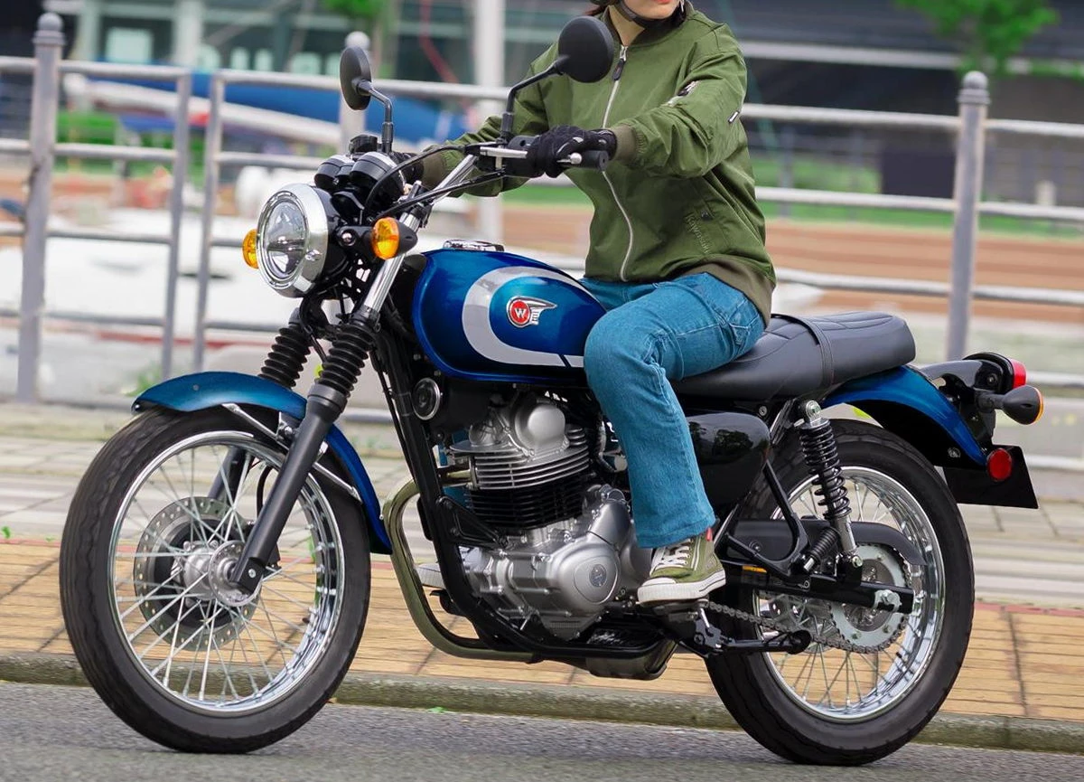 Une Kawasaki W 230 bleu, un motard en pantalon jean et blouson vert, des barrières