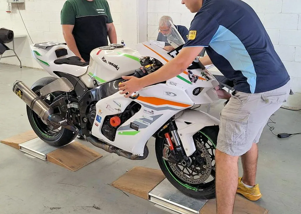 Une Kawasaki ZX-10R blanche dans un box, tenue par une homme en short gris et t-shirt bleu, un homme en t-shirt vert et noir.