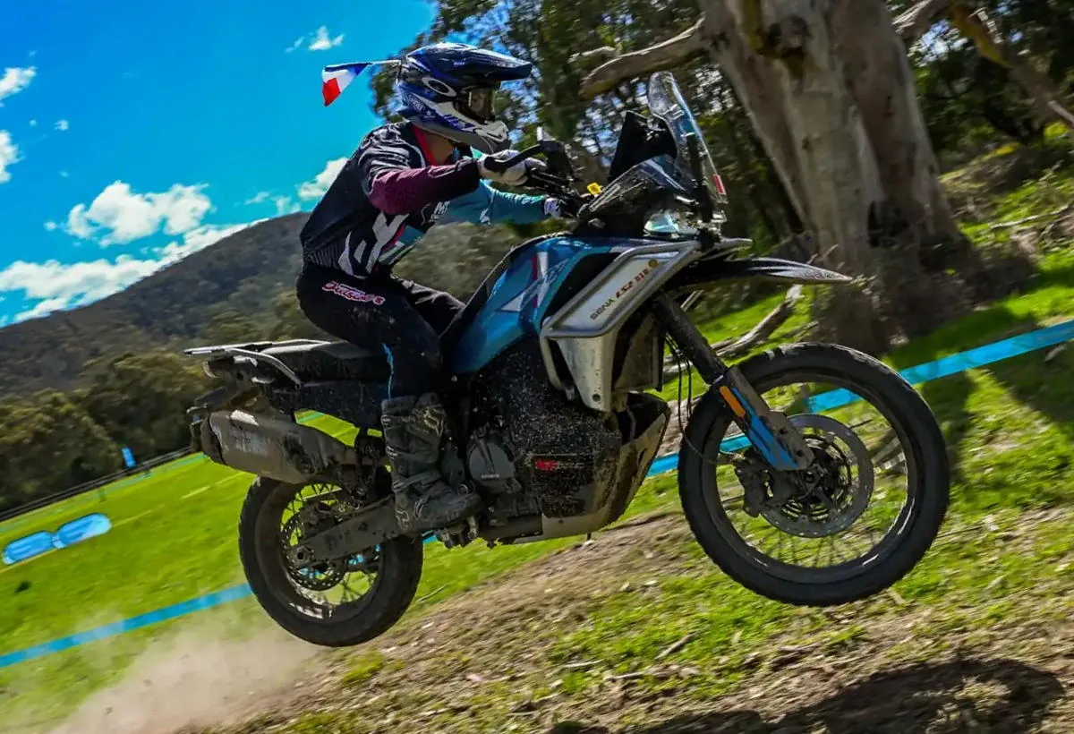 Une moto CFMOTO 800MT-X en plein saut, de l'herbe, un arbre, une colline et un ciel bleu