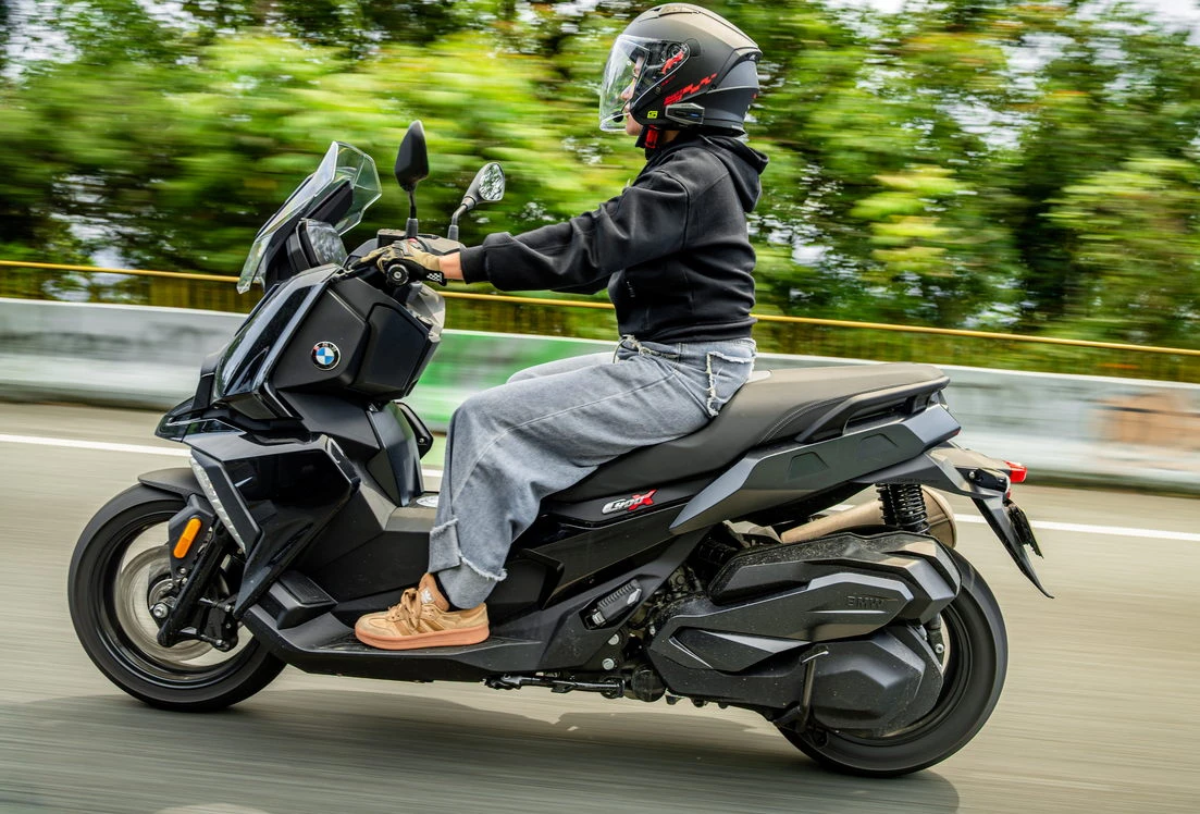 Un scooter BMW, une route, des arbres, une motarde avec un haut noir et un casque jet équipé d'un intercom Sena.