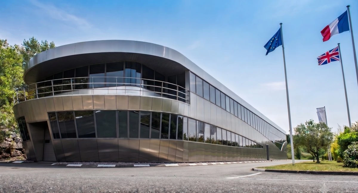 L'usine de Brough Superior à Toulouse, un bâtiment allongé avec beaucoup de vitres, un ciel bleu, les drapeaux européen, français et anglais.