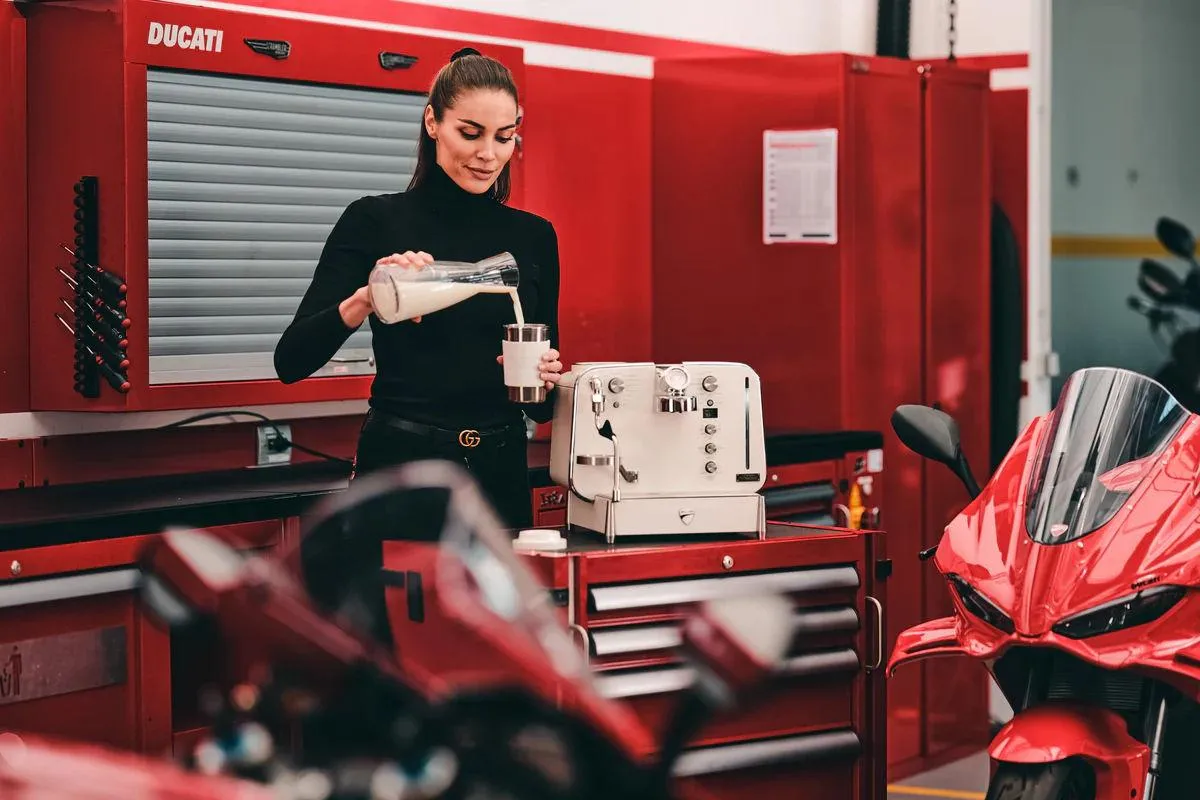 Une femme se prépare un café dans un garage, avec des ducati Rouge, des outils, et une machine barista blanche.