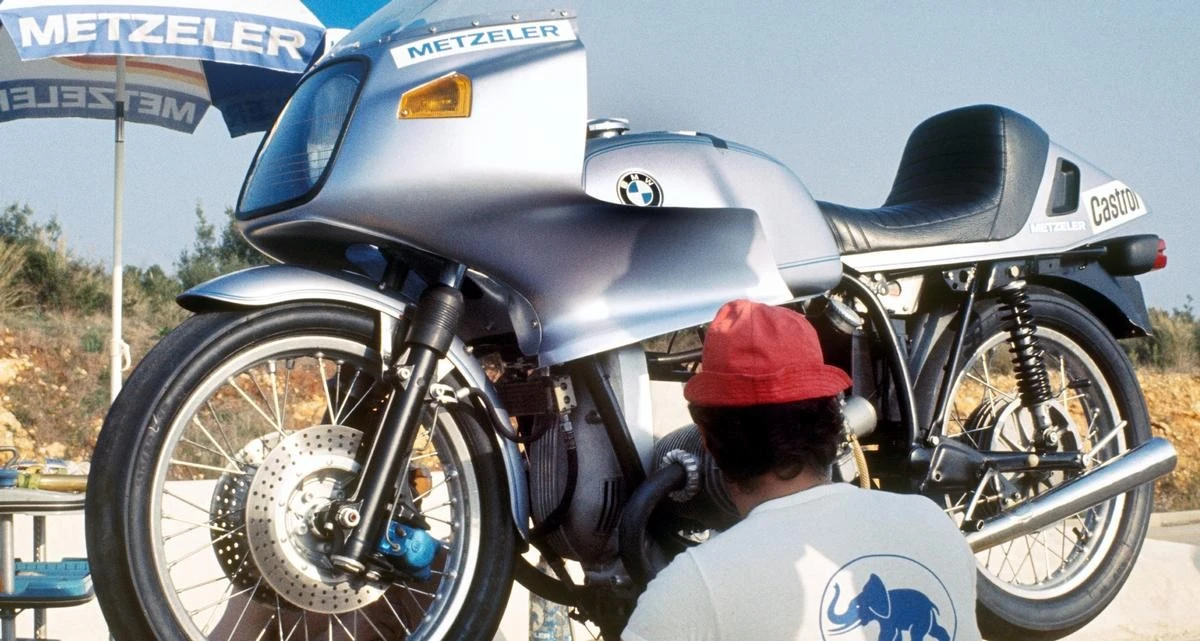 Une BMW R 100 RS grise sur un étable et un homme avec un t-shirt blanc Metzeler et une casquette rouge