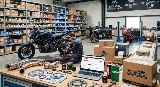 Image illustrant un comparatif des meilleurs sites de pièces moto en 2026. On y voit un atelier avec un mécanicien travaillant sur un roadster, entouré de pièces détachées neuves et d'occasion, ainsi que des colis prêts pour la livraison. Un ordinateur portable affiche une boutique en ligne spécialisée.