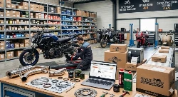 Image illustrant un comparatif des meilleurs sites de pièces moto en 2026. On y voit un atelier avec un mécanicien travaillant sur un roadster, entouré de pièces détachées neuves et d'occasion, ainsi que des colis prêts pour la livraison. Un ordinateur portable affiche une boutique en ligne spécialisée.