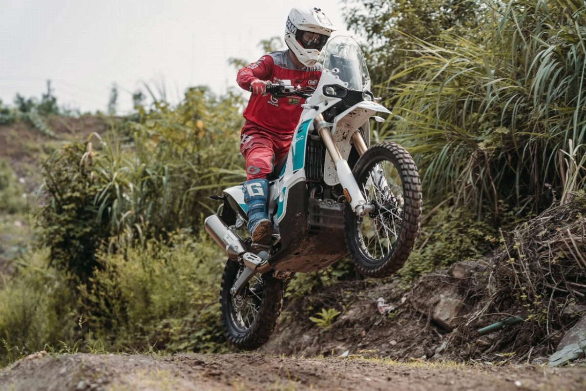 Pilote en combinaison rouge et casque blanc effectuant un saut en motocross sur une Kove 450 Rally blanche et bleue dans un décor de forêt
