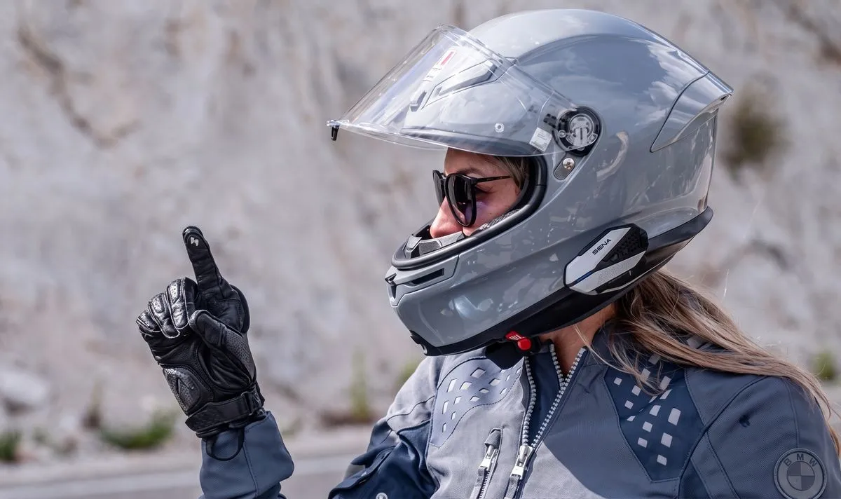 Une motarde avec un casque gris et le doigt levé dans un gant noir.
