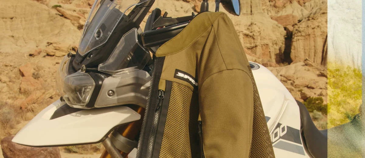 Un blouson moto kaki, l'avant d'une moto Triumph Tiger 900 et des rochers