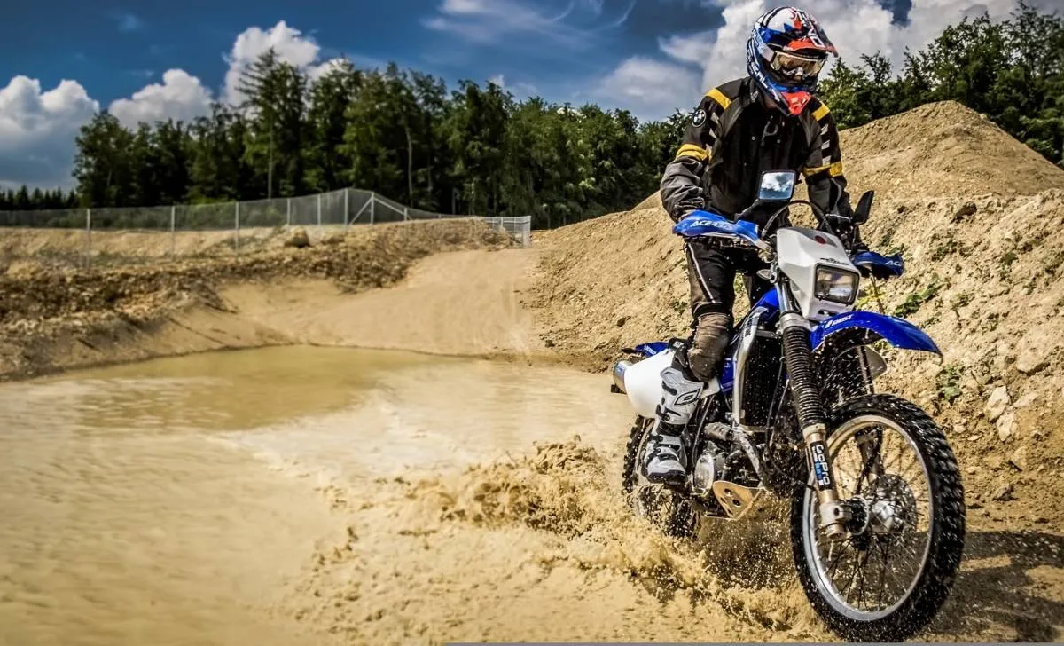 Une moto d'enduro bleu sortant d'une flaque d'eau et de boue, des arbres et un ciel bleu