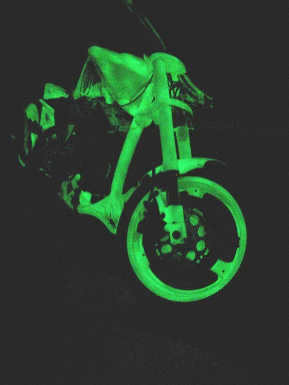 Une moto dans la nuit à laquelle on a appliqué une peinture phosporescente sur le cadre, les roues, le réservoir. Elle devient verte la nuit
