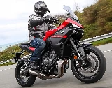 Une moto Yamaha Tracer 7 rouge et noire, une route, une barrière de sécurité, des herbes hautes, un fond de mer et un pilote habillé en vêtements gris