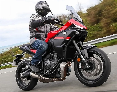 A red and black Yamaha Tracer 7 motorcycle, a road, a guardrail, tall grass, a seabed, and a rider wearing gray clothing
