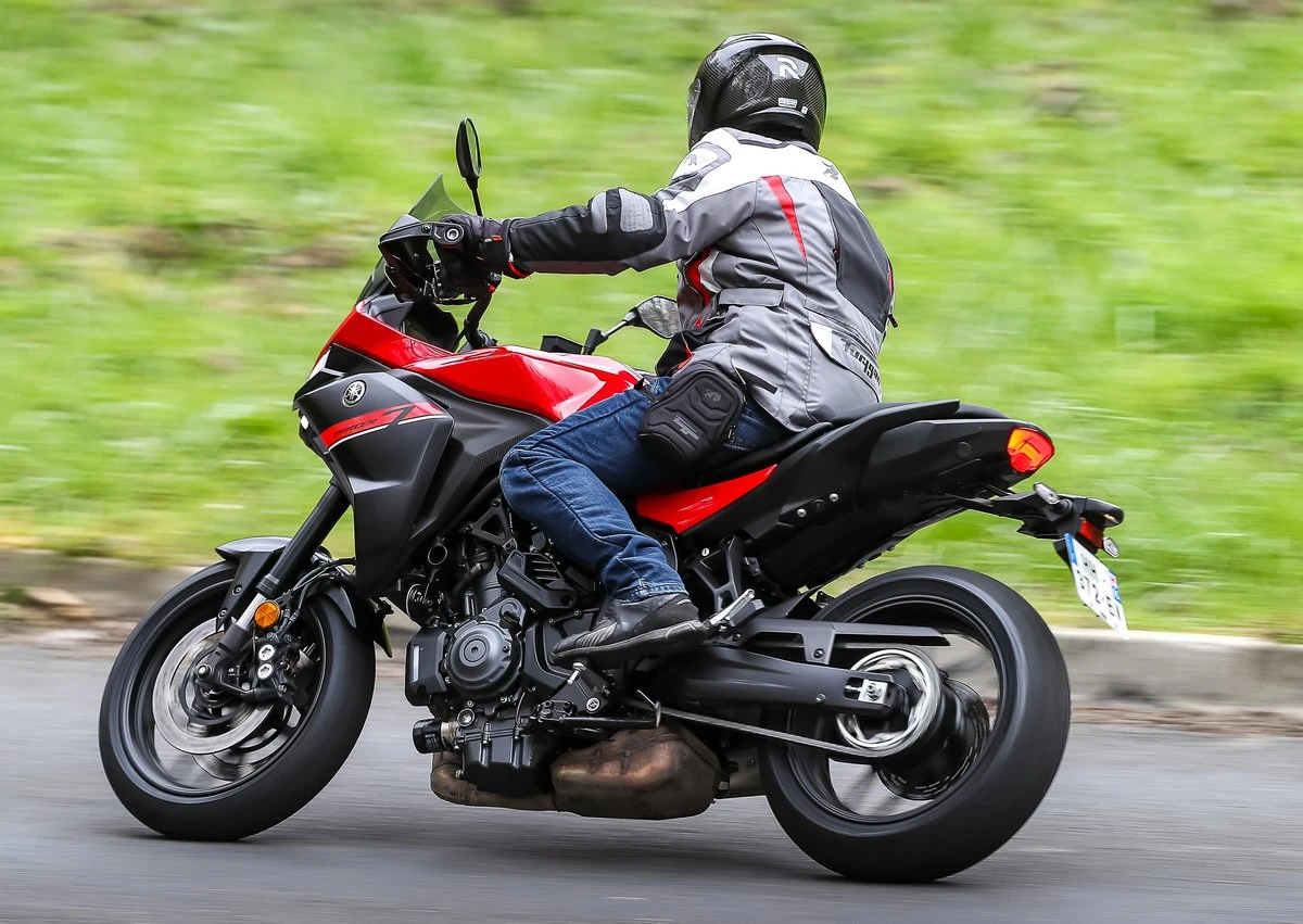 Une moto Yamaha Tracer 7 rouge et noire, une route, une barrière de sécurité, des herbes hautes, un fond de mer et un pilote habillé en vêtements gris