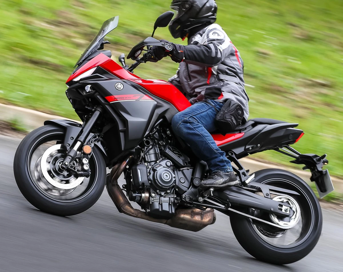 Une moto Yamaha Tracer 7 rouge et noire, une route, une barrière de sécurité, des herbes hautes, un fond de mer et un pilote habillé en vêtements gris